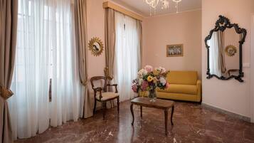 Ein Wohnzimmer mit einer Couch, einem Sessel, einem Tisch mit Blumenvase, einer Chandelüster und zwei Wandspiegeln.