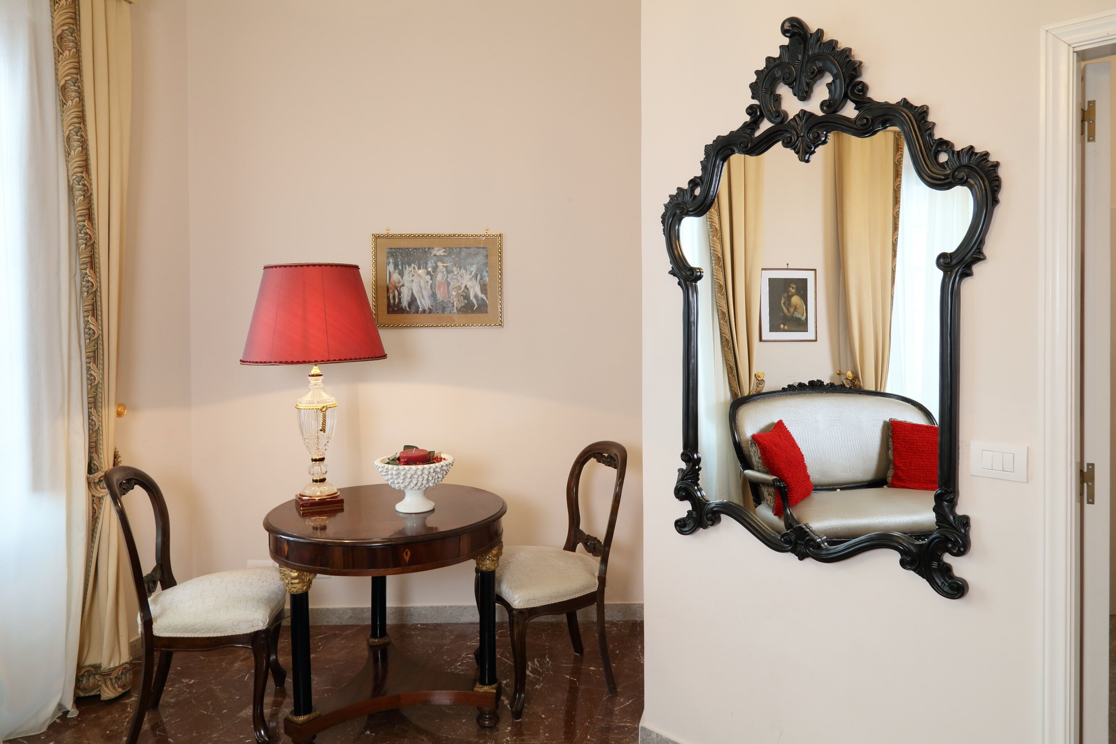 A room with a round table, two chairs, a red lamp, a mirror, and two framed pictures on the wall.