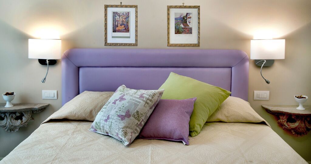 Una cama con cabecera morada, dos lámparas fijadas a la pared y dos cuadros encolados encima.