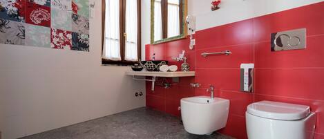 Un baño con pared de azulejos rojos, inodoro blanco, bidé y lavabo con espejo superior.