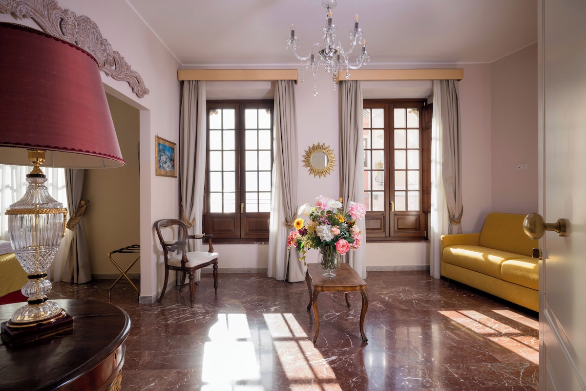 A room with a chandelier, a sofa, a table with a vase of flowers, and a lamp.