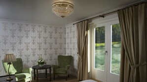 A room with a bed, two armchairs, a small table with a lamp, a chandelier, and a view of a garden through a door. - Hotel Hirvihaaran Kartano (Maentsaelae)