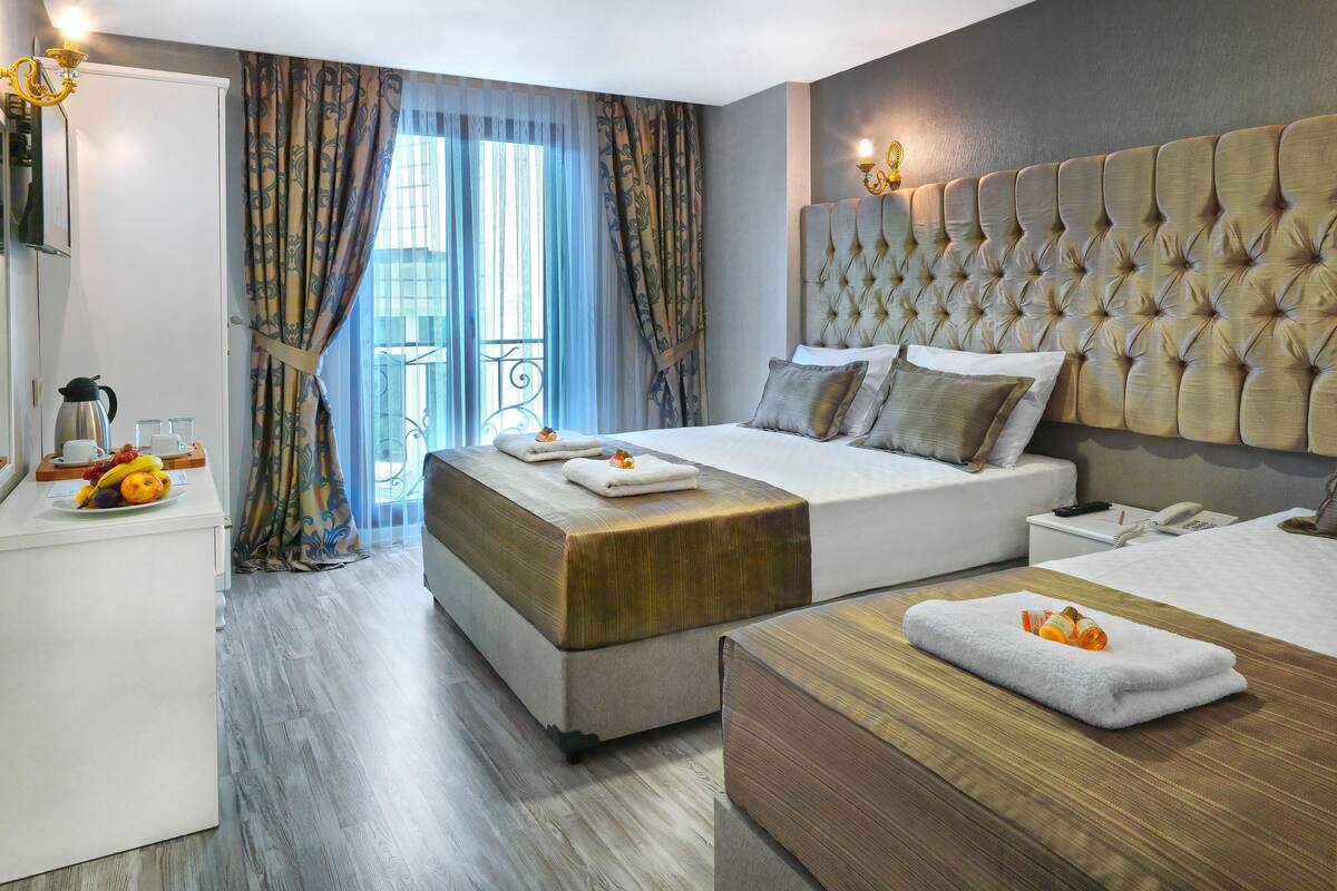 A hotel room with a large bed, a bedside table with a phone and remote, a bedside table with fruit and a coffee maker, and a view of the outside through a window with curtains.