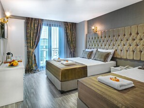 Habitación de hotel con una cama grande, una mesita de noche con teléfono y control remoto, otra mesita de noche con fruta y cafetera, y vistas al exterior a través de una ventana con cortinas.