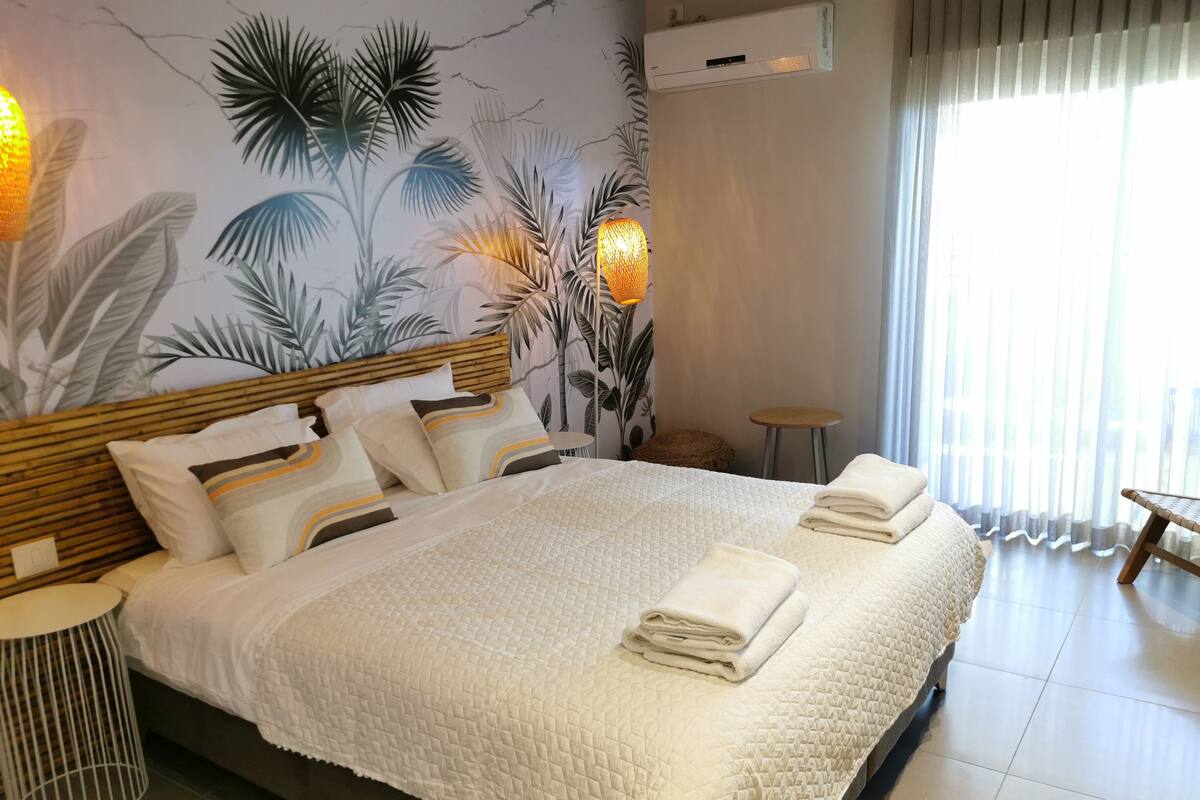 A modern hotel room with a large bed, a wall mural of tropical leaves, a small round table, and a window with curtains.
