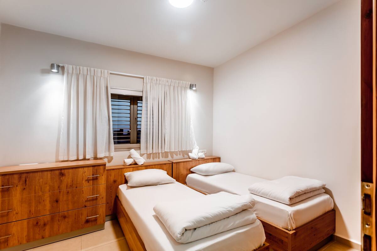 A bedroom with two single beds, a wooden dresser, a mirror, and a window with curtains.