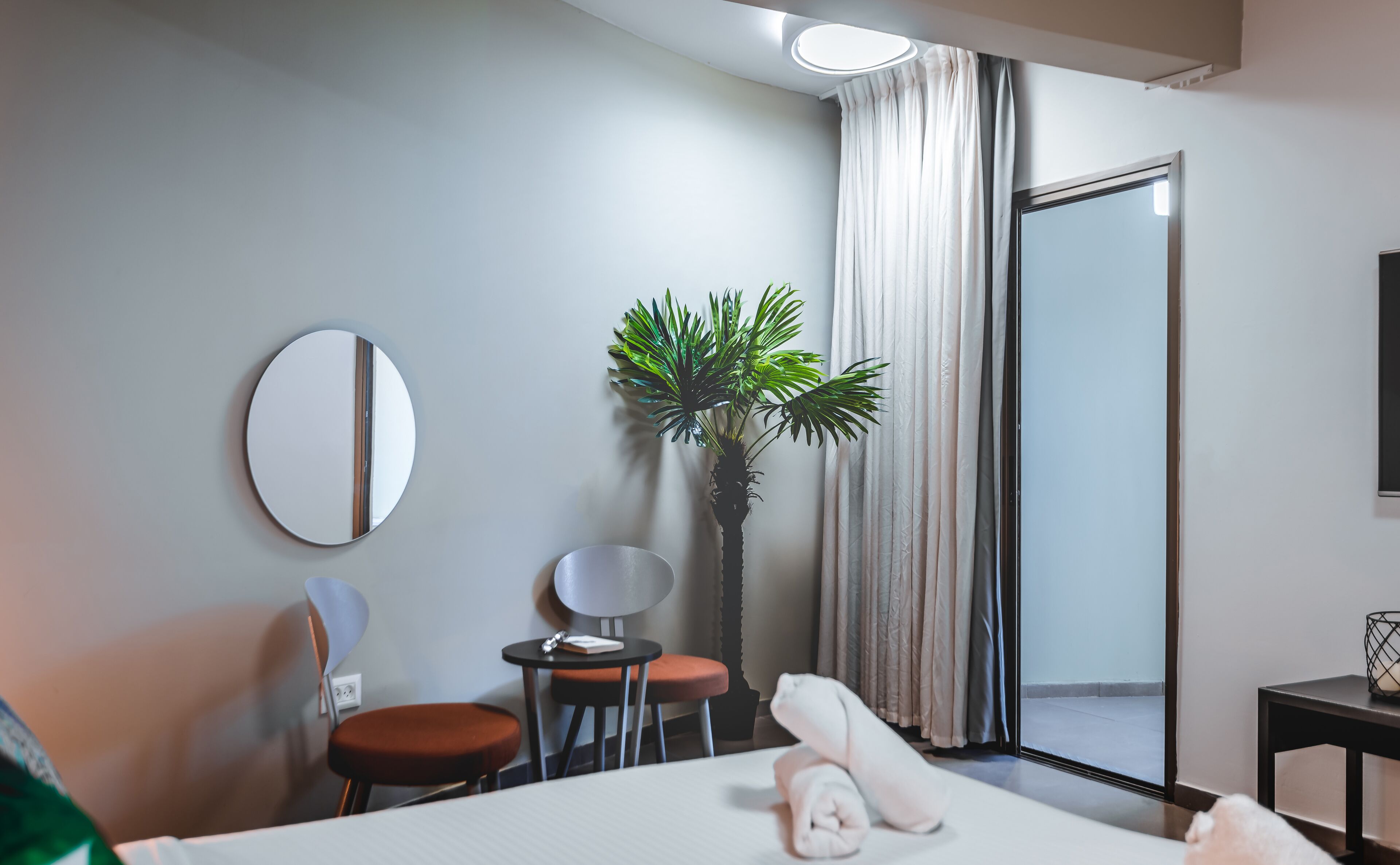 Ein Hotelzimmer mit einem Bett, einem runden Spiegel, einem kleinen Tisch mit Stuhl, einer Topfpflanze und Blick ins Freie durch ein großes Fenster.