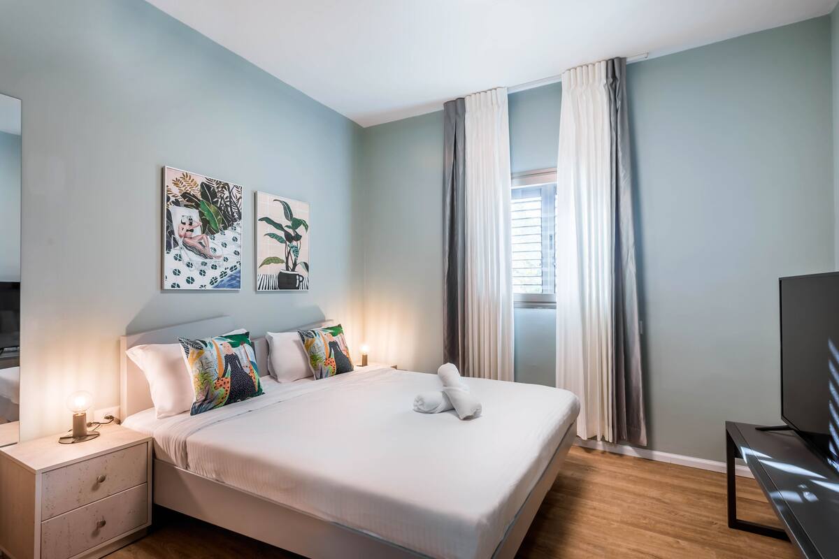 A neatly made bed with white linens and a folded towel, a bedside table with a lamp, and a TV on a stand.