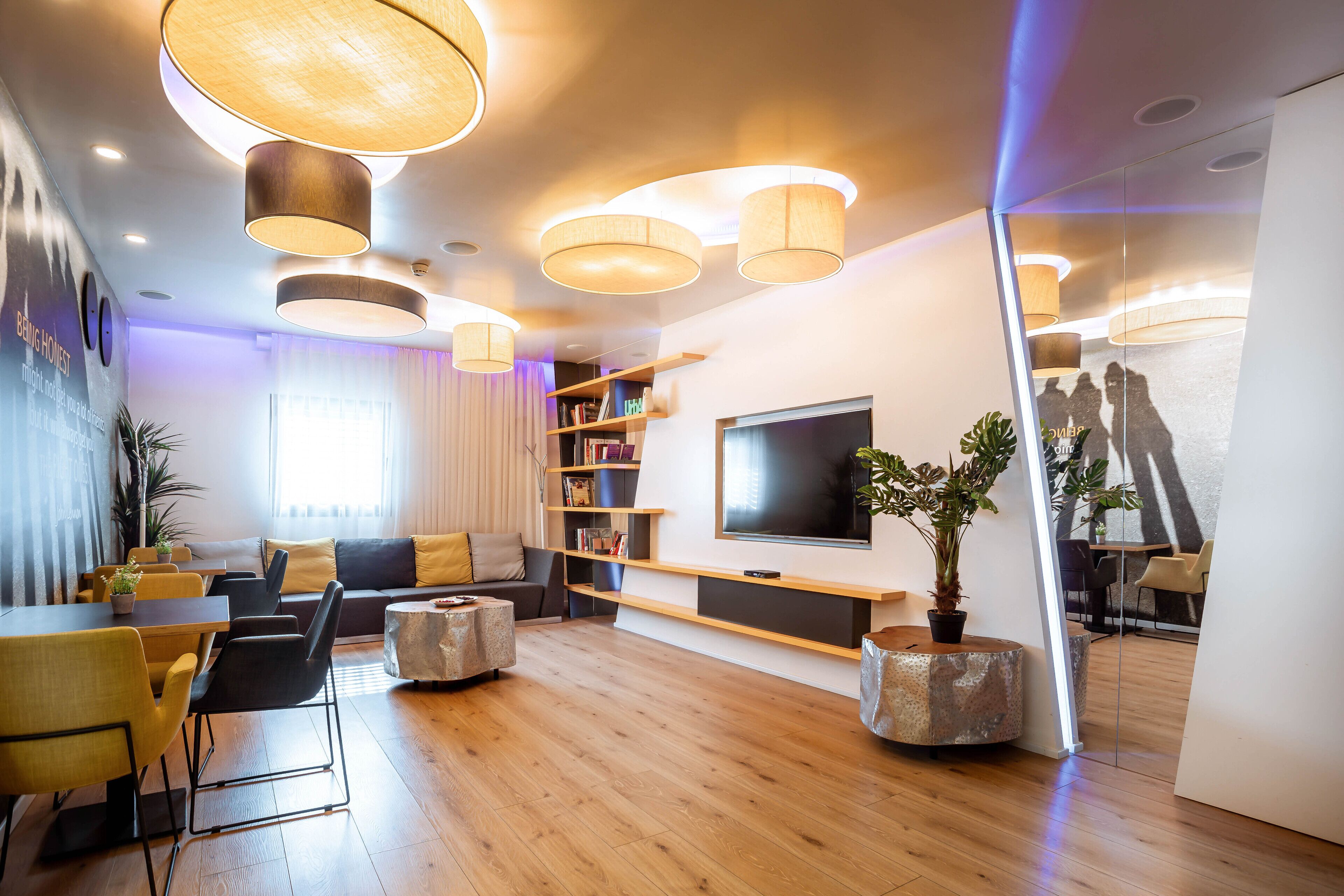 A modern living room with a wooden floor, a sofa, a dining table with chairs, a TV mounted on the wall, and a bookshelf.