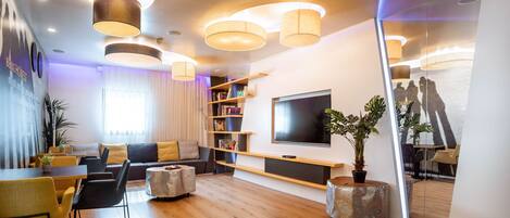 A modern living room with a wooden floor, a sofa, a dining table with chairs, a TV mounted on the wall, and a bookshelf.