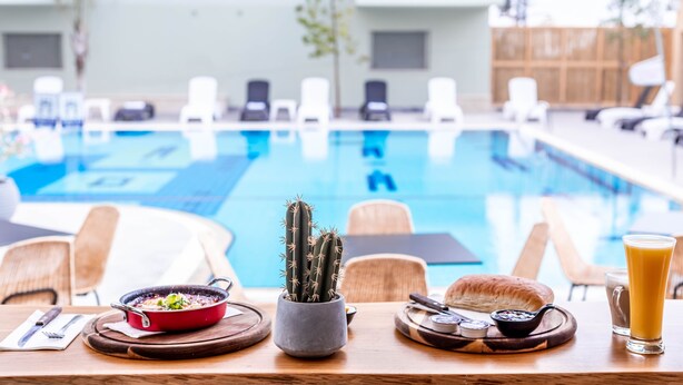 A dining table with a red pot, a cactus, bread, and condiments. In the background, there is a pool with lounge chairs. - Almog Eilat (Eilat)