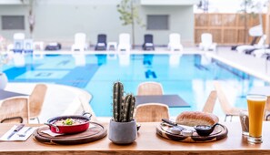 A dining table with a red pot, a cactus, bread, and condiments. In the background, there is a pool with lounge chairs. - Almog Eilat (Eilat)