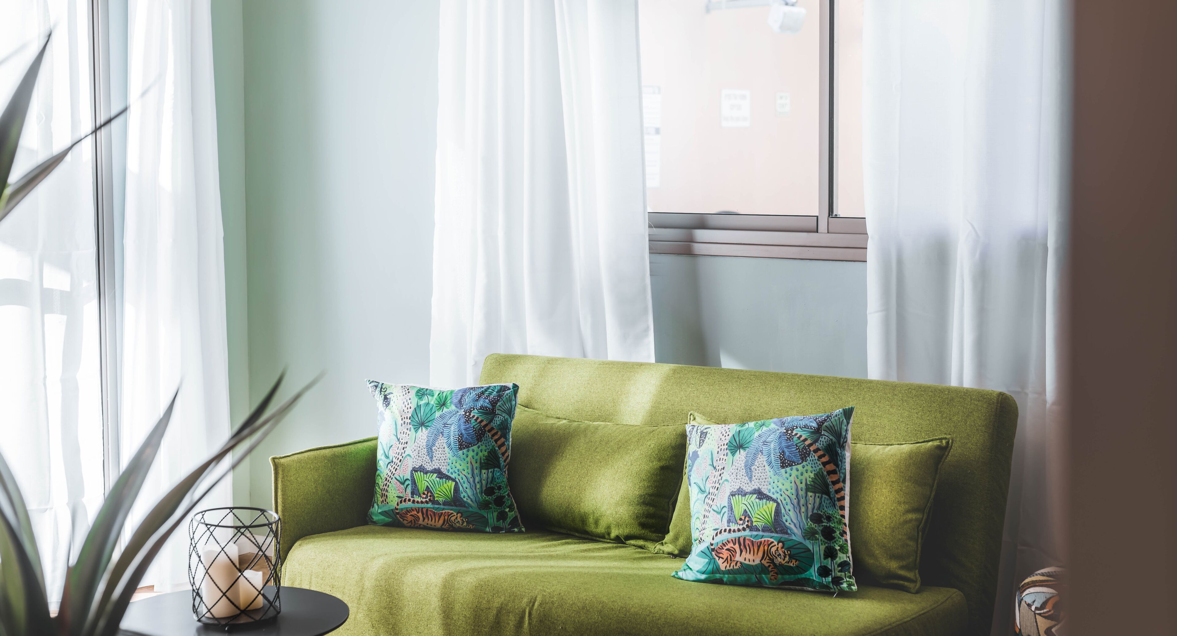 A green sofa with two patterned cushions, a small table with a candle, and a plant.