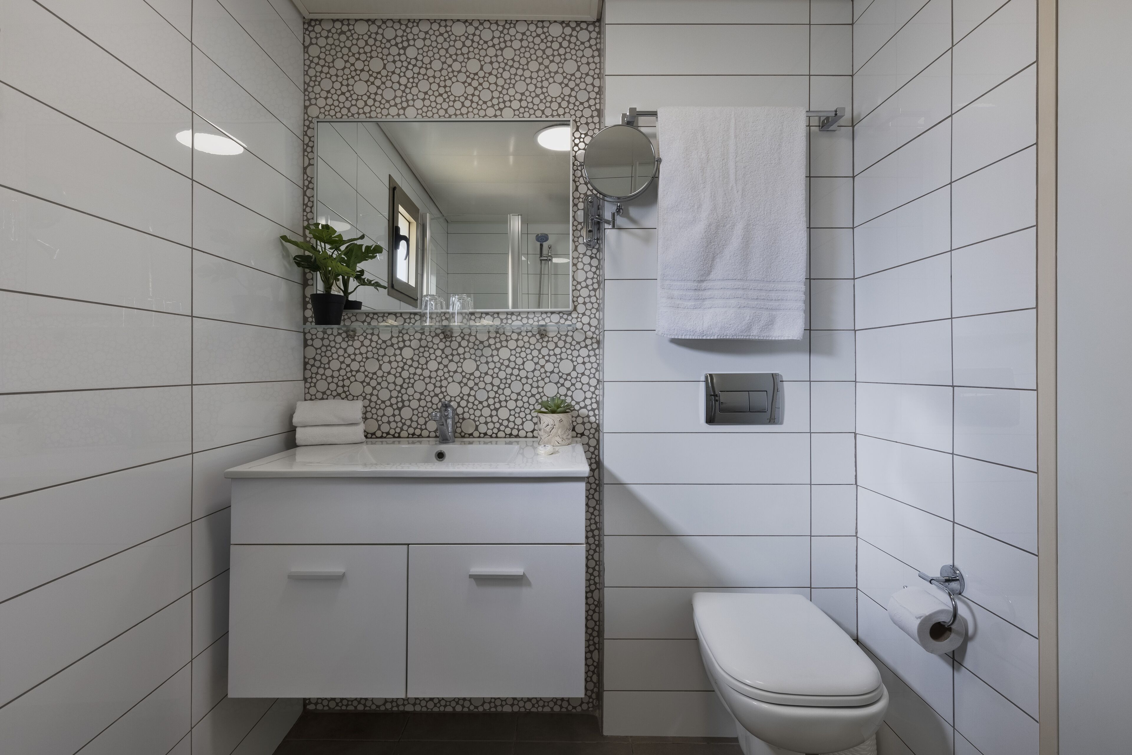 Ein Badezimmer mit weißen Fliesen, einem Waschbecken mit Spiegel, einer Toilette und einem Handtuch an der Wand.