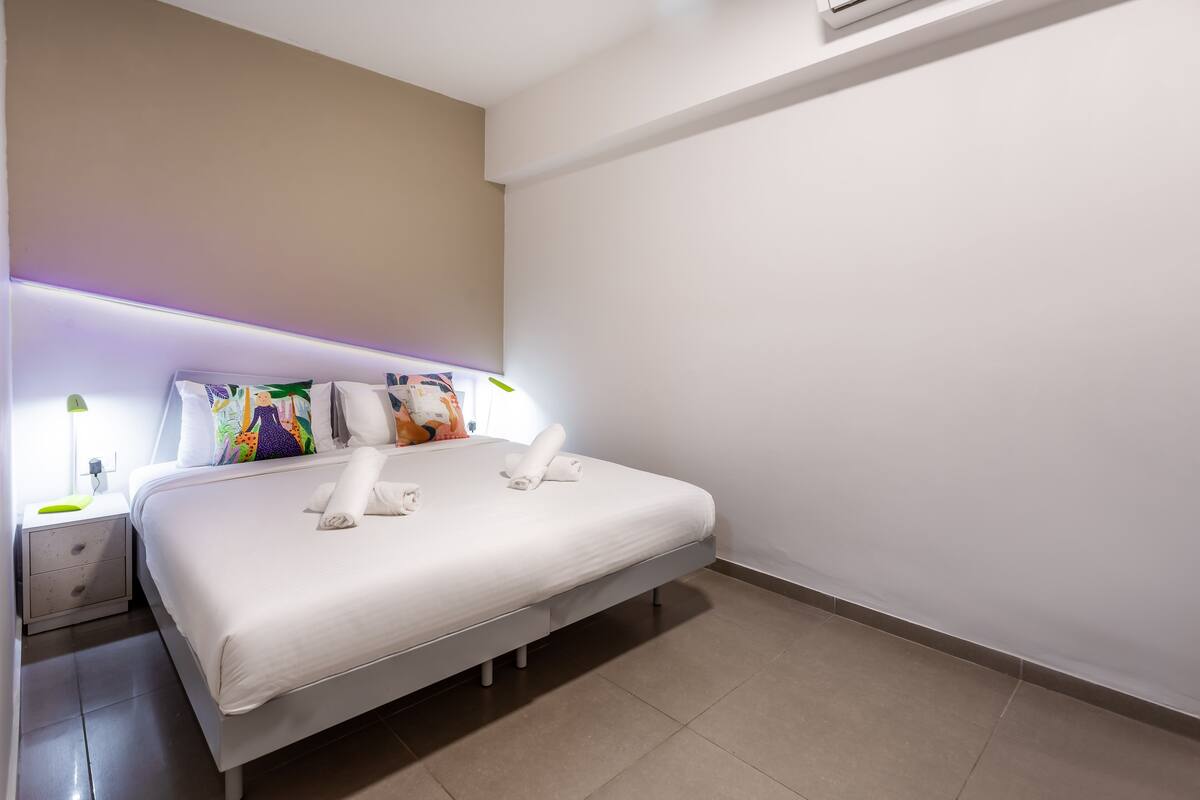 A modern hotel room with a neatly made bed, a bedside table with a lamp, and a plain wall.