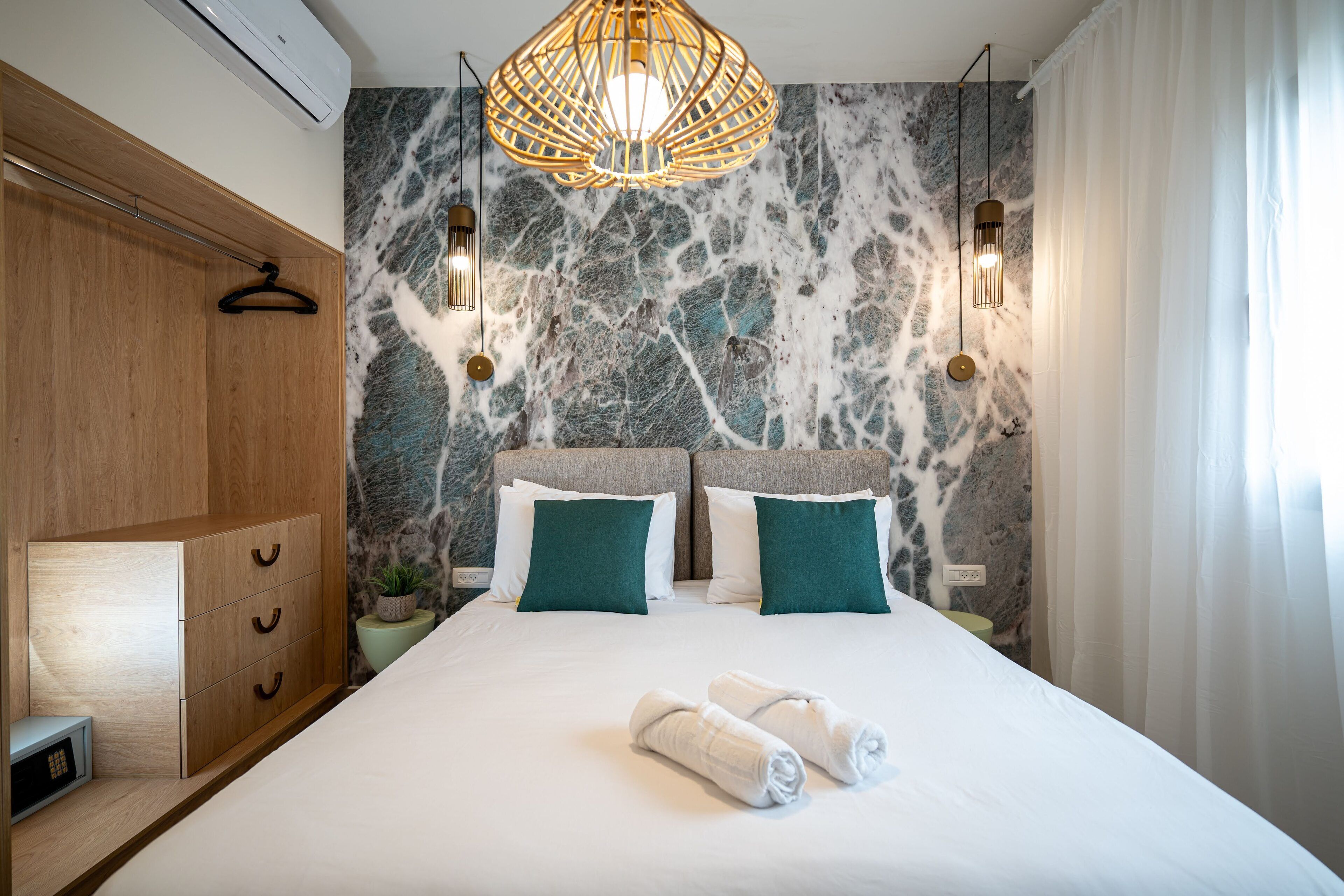 A hotel room with a bed, wooden wardrobe, marble wall, two bedside lamps, and a hanging light fixture.