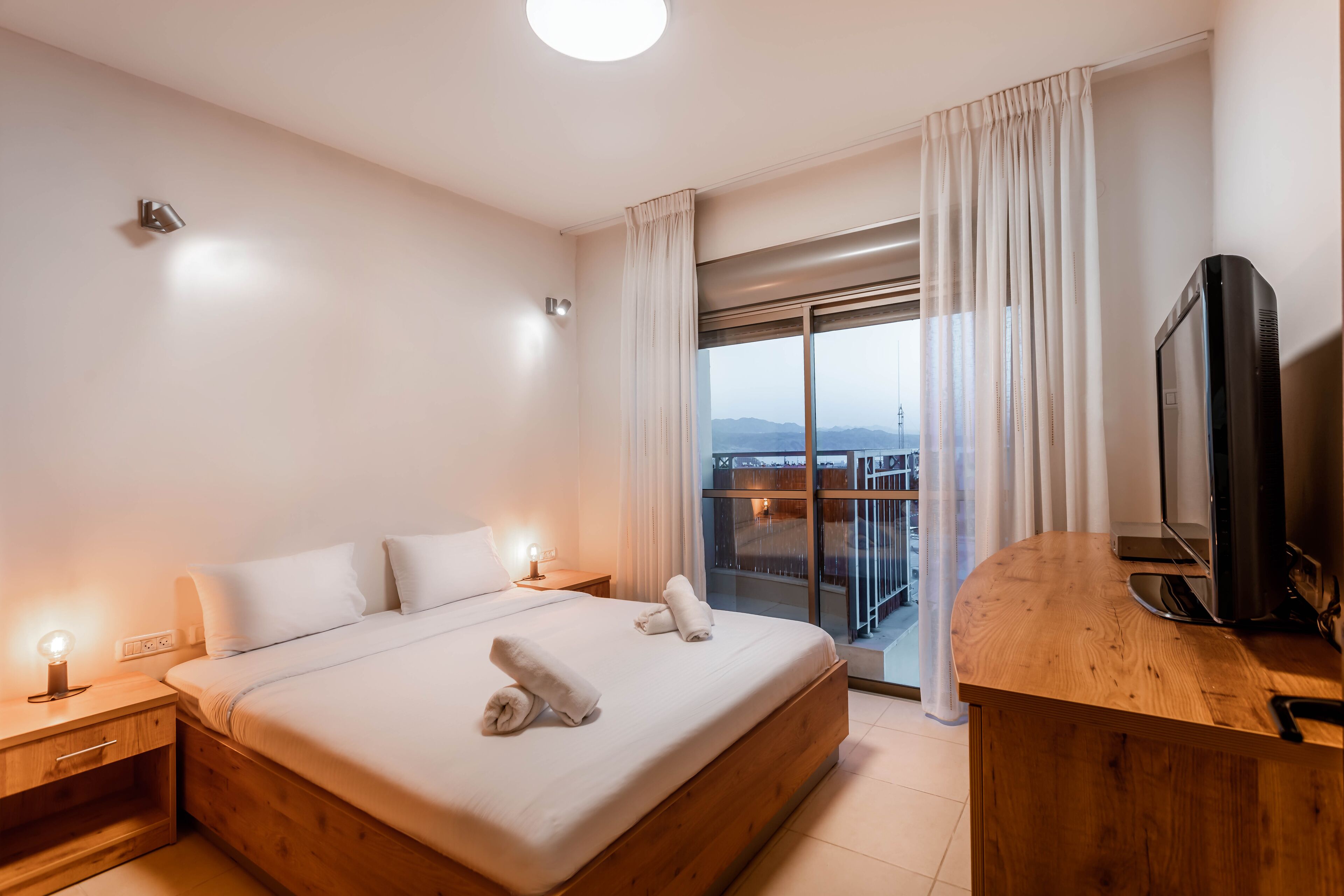 Moderni hotellihuone, jossa on suuri sänky, puinen työpöytä, taulutelevisio ja parveke, jolta on näkymä kaupunkiin.