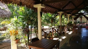 Un espacio al aire libre cubierto con mesas y sillas de madera, rodeado de exuberante follaje tropical y un techo de paja.