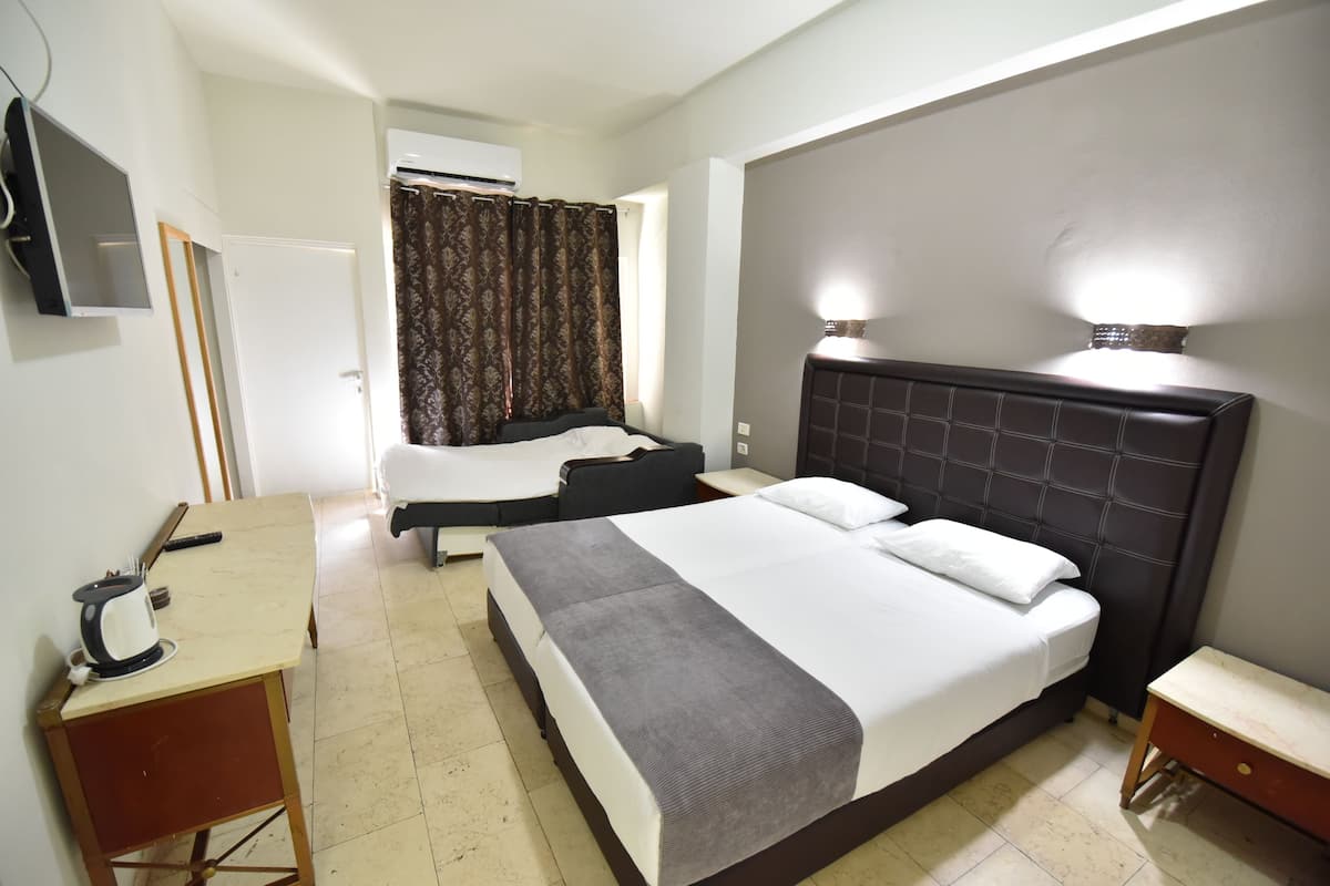 A hotel room with a large bed, a desk with a kettle, a television, and an air conditioning unit.