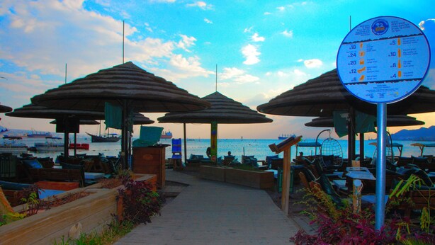 A seaside walkway with umbrellas and seating areas, a signboard with information, and boats in the background. - Maman Hotel (Eilat)