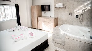 A hotel room with a bed, a wooden wardrobe, a TV mounted on the wall, a bathroom with a bathtub, and towels on the bed. - Maman Hotel (Eilat)