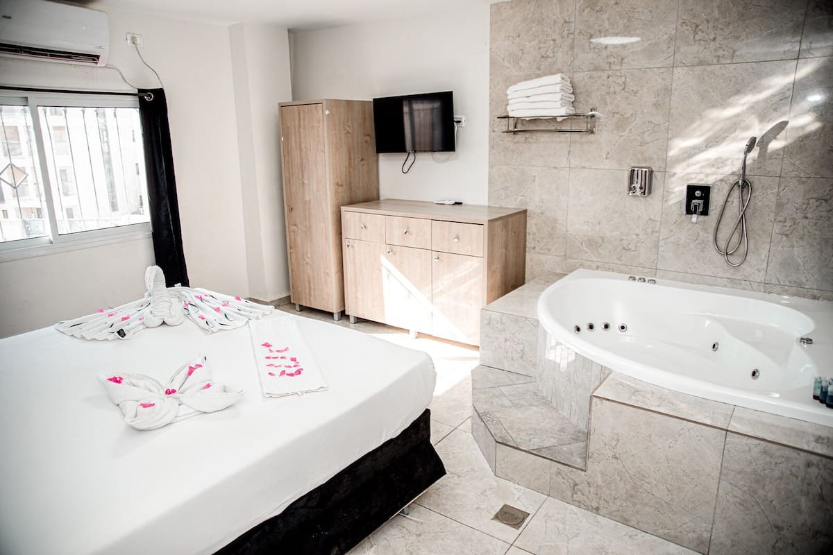 A hotel room with a bed, a wooden wardrobe, a TV mounted on the wall, a bathroom with a bathtub, and towels on the bed.
