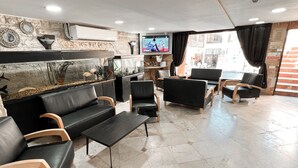 A modern lobby with black leather sofas, wooden chairs, a fish tank, and a TV on the wall. - Maman Hotel (Eilat)