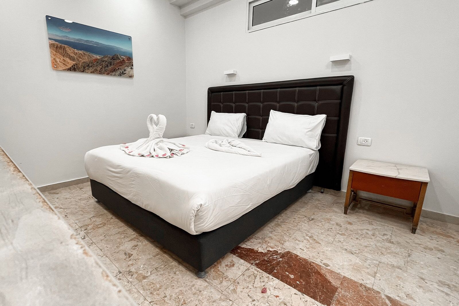 Een net opgemaakt bed met witte lakens en handdoeken, een donker hoofdbord, een klein nachtkastje en een schilderij aan de muur.