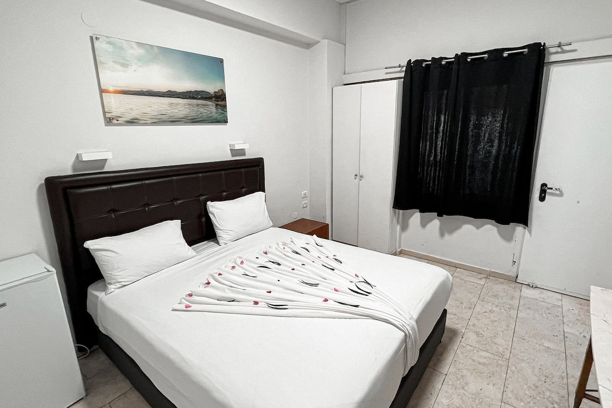 A neatly made bed with white linens and a floral pattern, a dark wooden headboard, a small bedside table, a white wardrobe, and a black curtain on a window.
