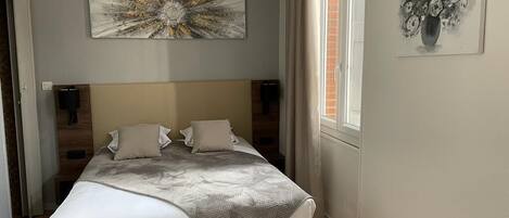 A bedroom with a bed, a headboard, a wall-mounted artwork, a window with curtains, and a chair.