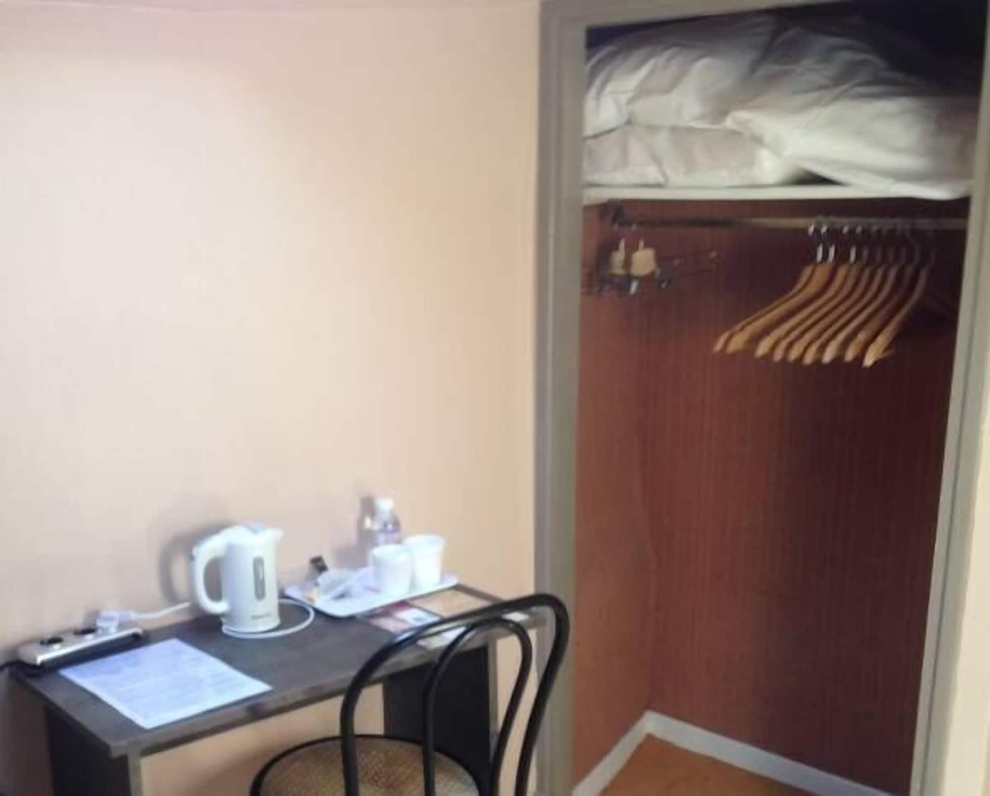A small room with a desk, a chair, a kettle, and a closet with hanging clothes.