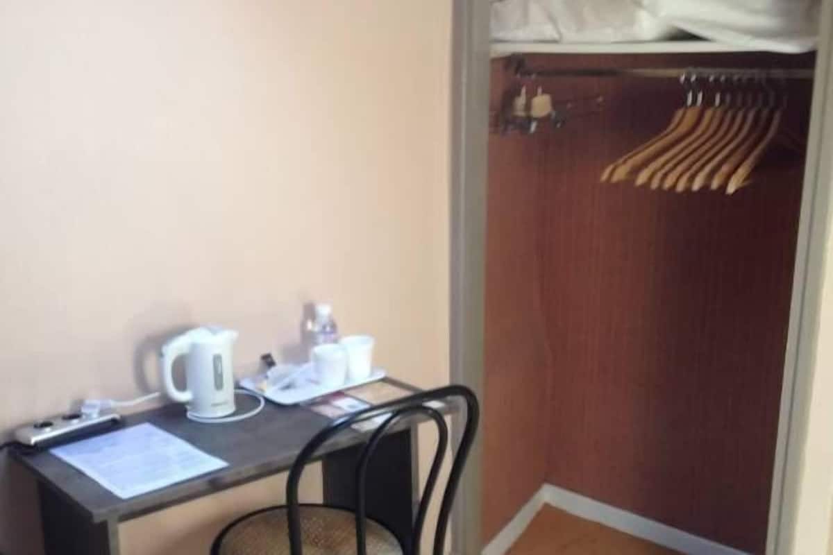 A small room with a desk, a chair, a kettle, and a closet with hanging clothes.