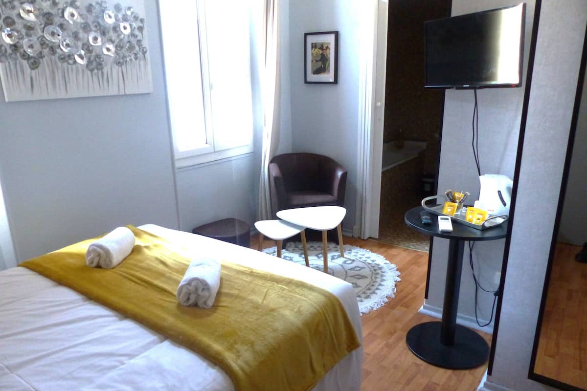 A hotel room with a bed, a small table, a chair, a TV, and a desk with a computer and phone.