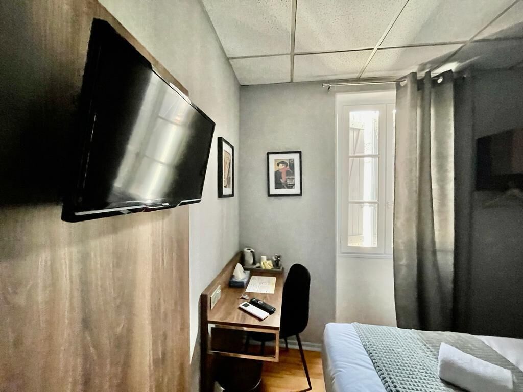 A compact hotel room with a bed, a desk, a chair, a TV mounted on the wall, and a window with curtains.
