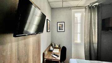 A compact hotel room with a bed, a desk, a chair, a TV mounted on the wall, and a window with curtains.