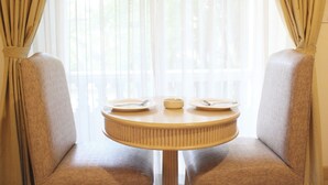 A round table with two plates and a bowl, set between two cushioned chairs, with sheer curtains and a view of greenery outside. - Romance Hotel Srinakarin (Bangkok)