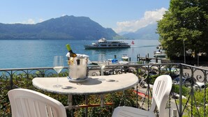 Terraza junto al lago con una mesa dispuesta para comer, con vistas a un lago tranquilo con barcos y montañas al fondo.
