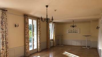 A spacious room with wooden flooring, large windows with floral curtains, and a chandelier.