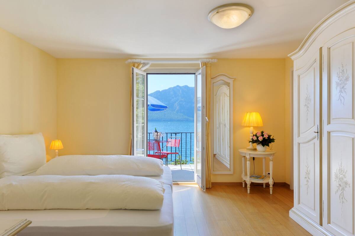 Une chambre d’hôtel avec un lit, une table de chevet avec une lampe, une petite table avec une lampe et des fleurs, et une vue sur la mer par une porte ouverte.