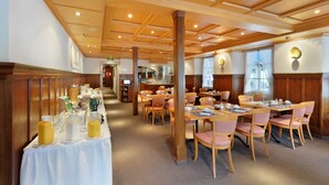 Comedor con paneles de madera, mesas dispuestas con sillas rosadas y una barra buffet con comida y bebidas.