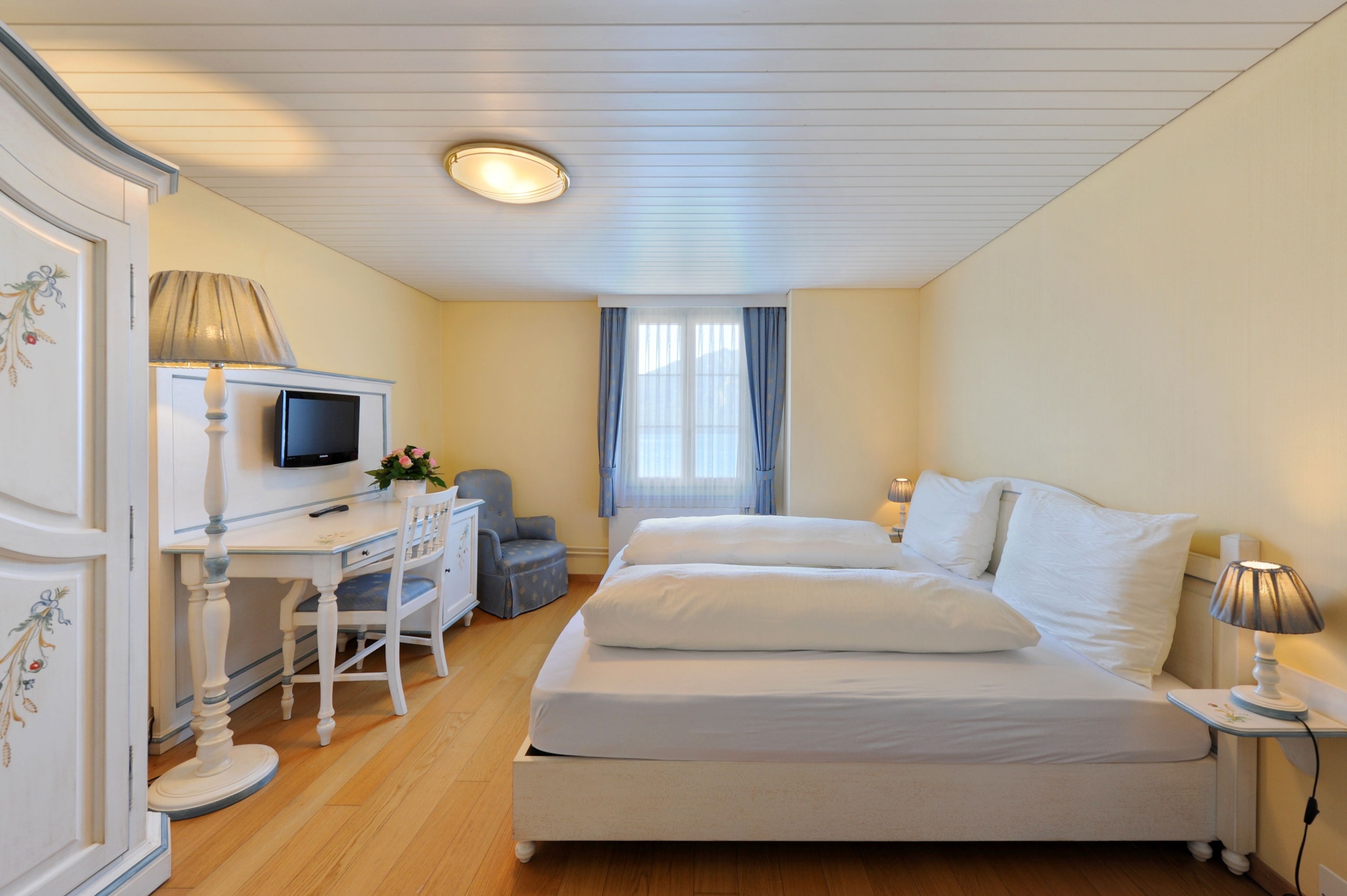 Habitación de hotel con cama, escritorio con silla, televisor y ventana con cortinas.
