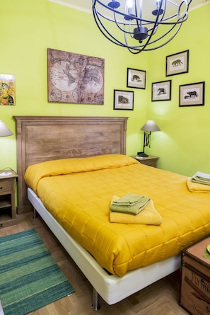 A neatly made bed with a yellow quilt, a green rug, a wooden headboard, and a wall with framed animal pictures. - La Quercia di Mamre (Rome)