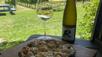 En pizza på en träbricka, tillsammans med en vinflaska och ett glas vitt vin, står på ett bord med utsikt över en vingård och berg i bakgrunden.