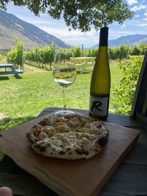 A pizza on a wooden board with a bottle of wine and a glass of white wine on a table with a view of a vineyard and mountains in the background.