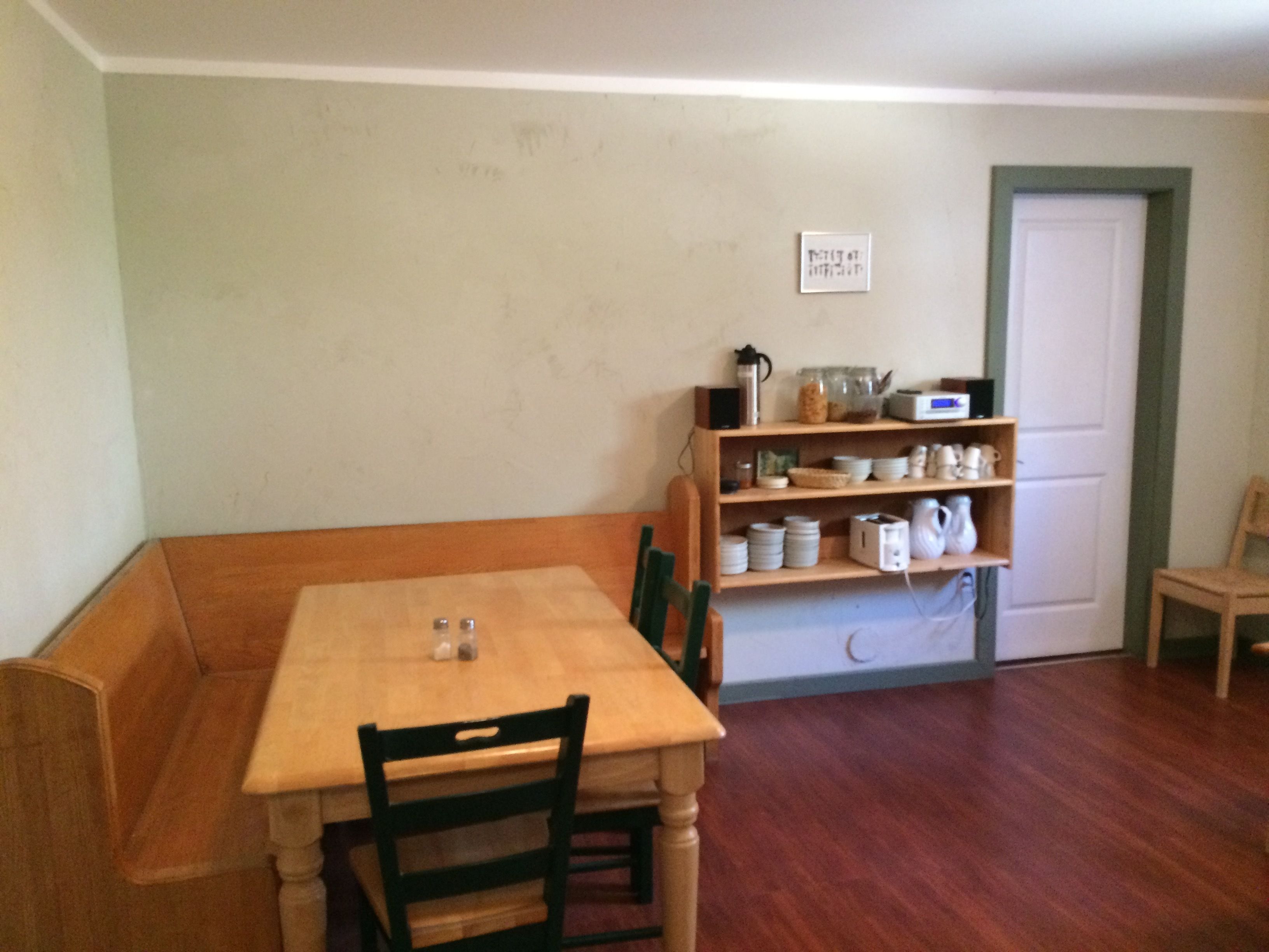 Una habitación pequeña y acogedora con una mesa y sillas de madera, un estante con utensilios de cocina y una puerta con marco verde.
