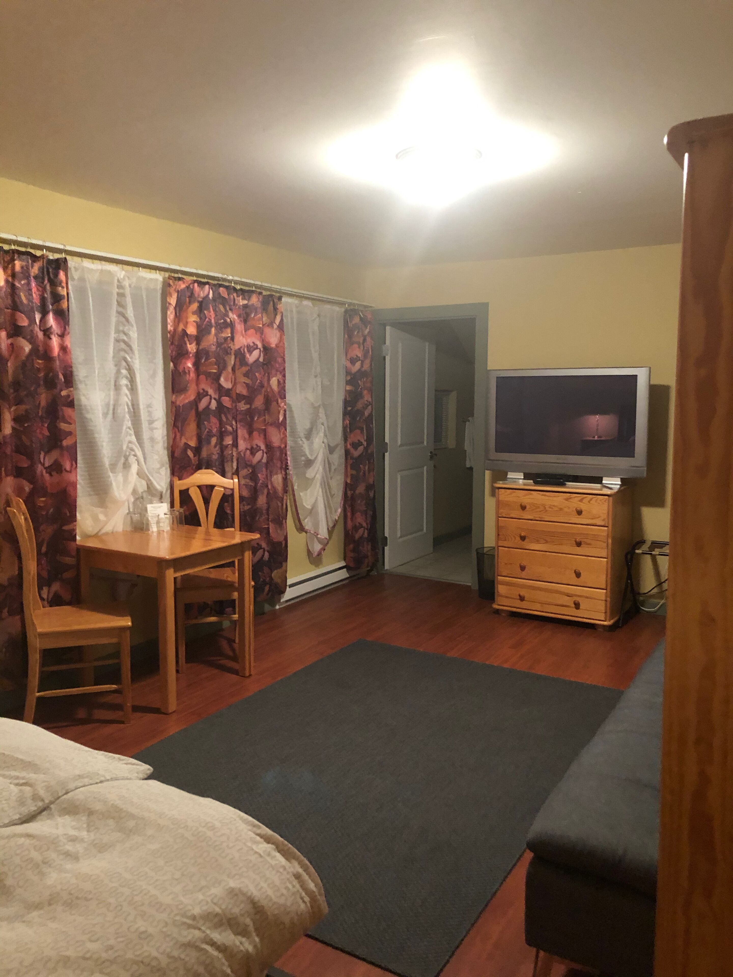 Una camera da letto con un letto, una cassettiera in legno, una televisione, una scrivania con una sedia e una porta che dà su un'altra stanza.