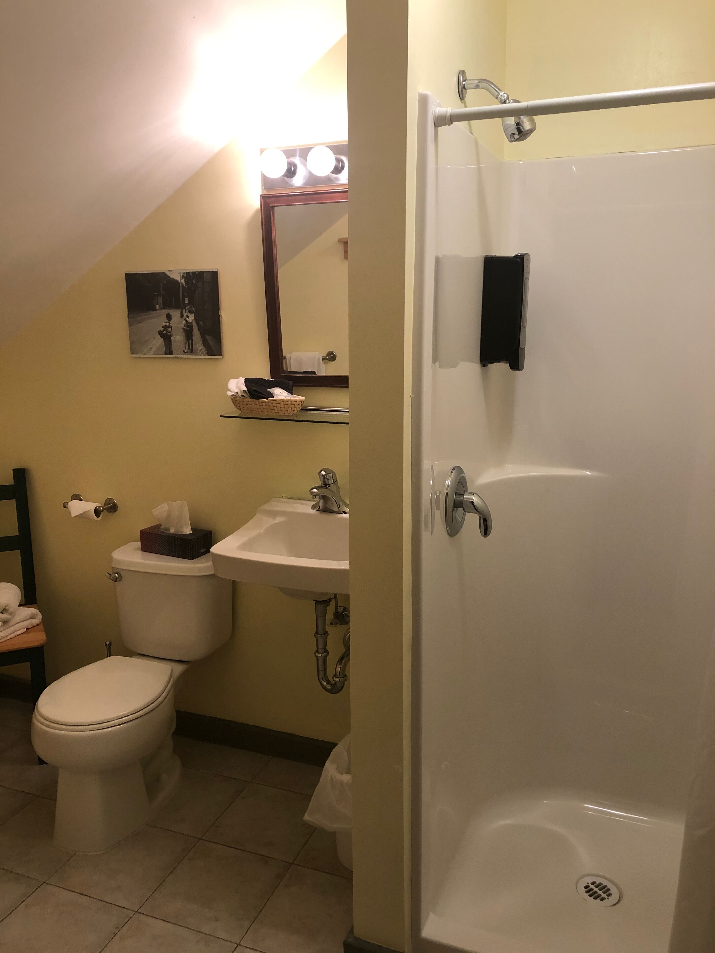 Un baño con inodoro, lavamanos y ducha. Hay un espejo con luces arriba y un perchero para toallas.