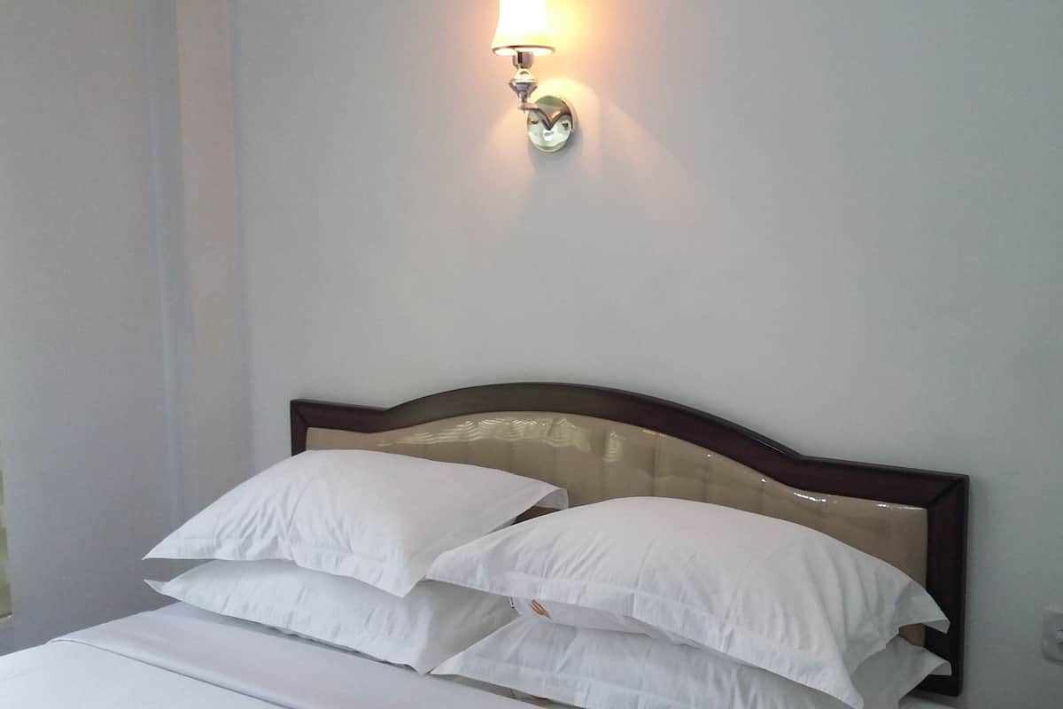 A neatly made bed with white linens and a wooden headboard, a wall-mounted light fixture, and a power outlet on the wall.
