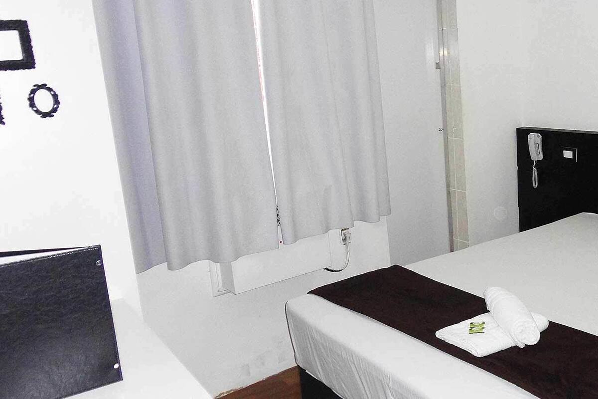A hotel room with a bed, white curtains, a black headboard, and a white pillow.