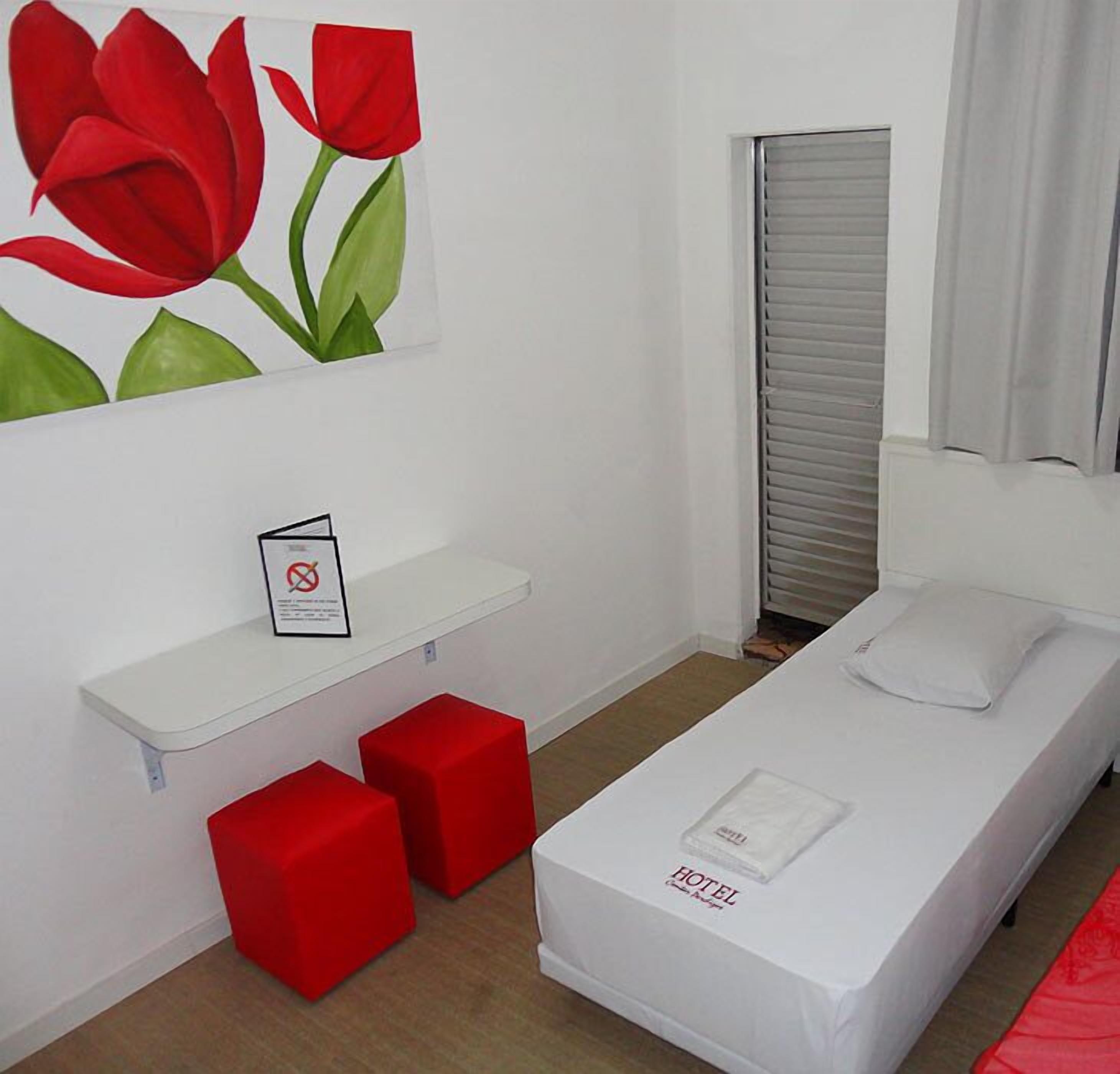 A single-bed room with a white bed, red ottomans, a red flower painting, a white shelf with a sign, and a closed sliding door.